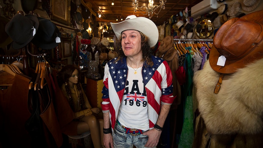 Mature-aged man with curly hair, a US jacket, and a cowboy hat smiles inside his small shop in Potts Point.