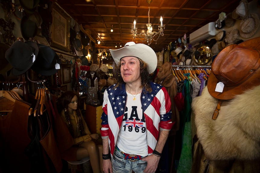 Mature-aged man with curly hair, a US jacket, and a cowboy hat smiles inside his small shop in Potts Point.