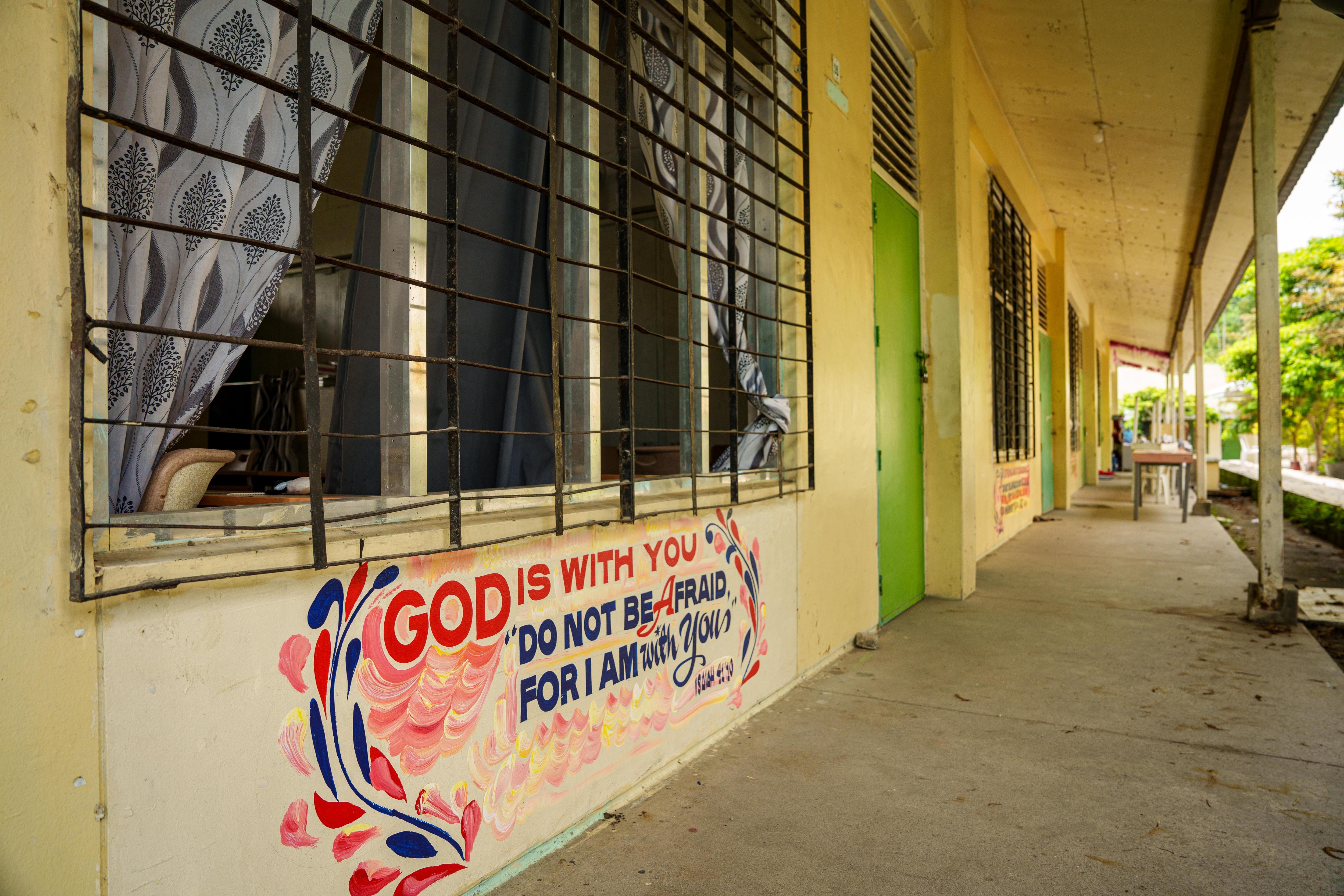The outdoor corridor of a classroom building. A bible verse is painted beneath the window of one.