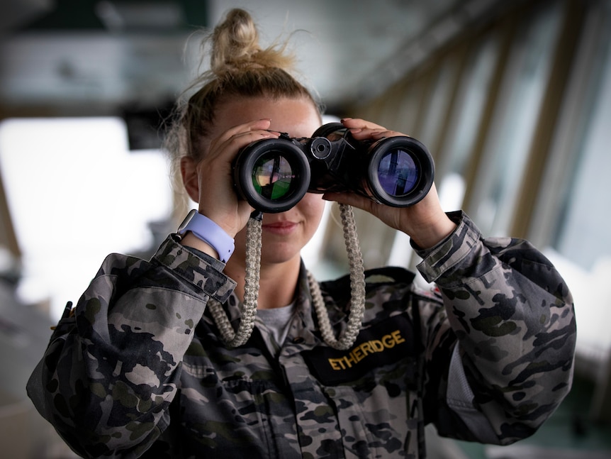 A woman in military uniform looks through a pair of binoculars on the deck of a ship.