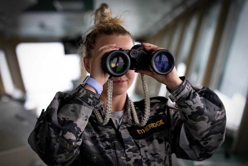 A woman in military uniform looks through a pair of binoculars on the deck of a ship.