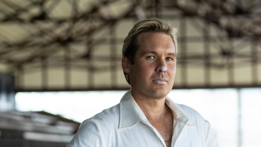 A man with white zinc on his face, blonde hair and cricket whites stands with an unsmiling gaze. 