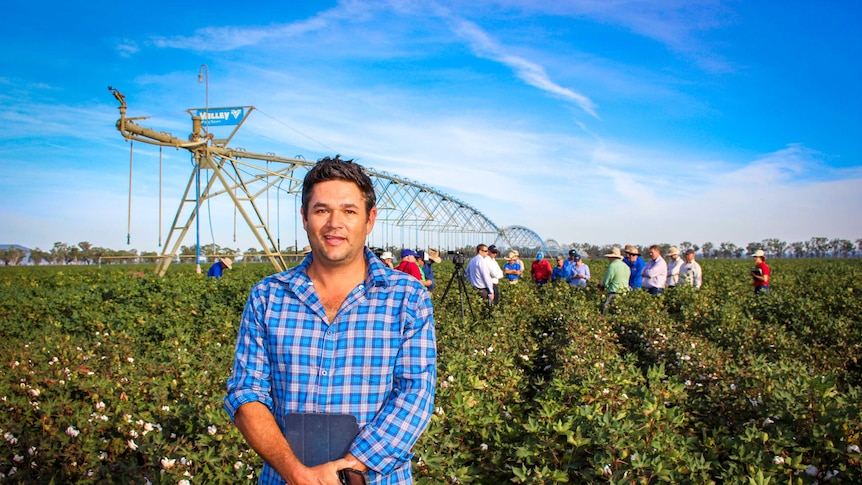 A man who looks to be in his 30s stands in a field wearing a bright, checked blue shirt. A group of people stands behind him.