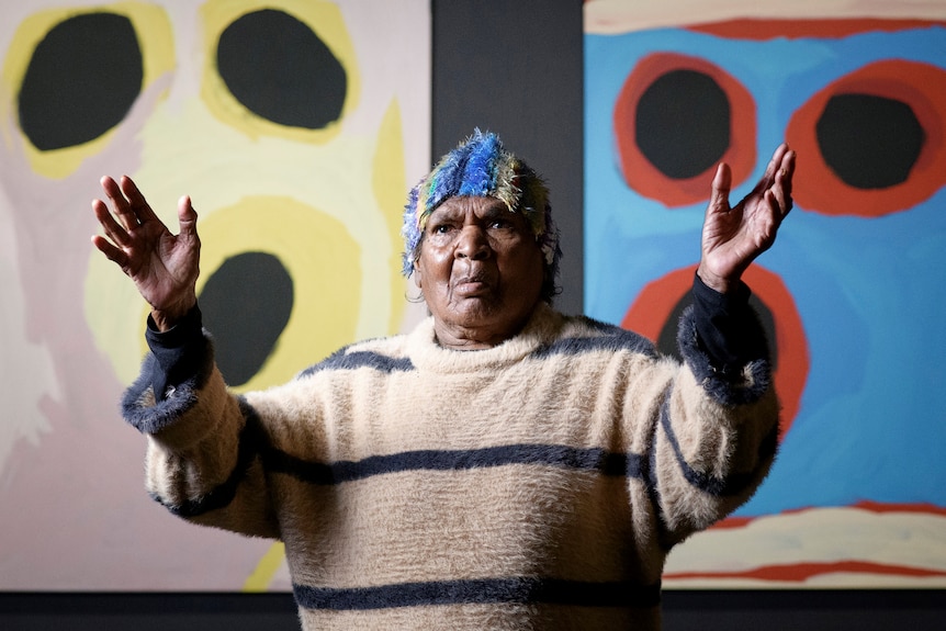 An older Aboriginal woman in jumper and beanie stands in front of bright artworks with her arms up