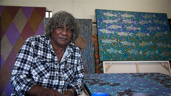 An artist stands in front of his colourful art works, including one with the Bundjalung 'diamond'
