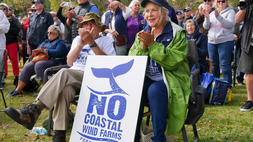 Two people sitting beside a placard that reads 'No coastal wind farms Port Stephens' with a crowd behind them