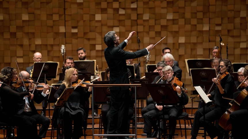 Conductor Eivind Aadland stands on a podum conducting the orchestra