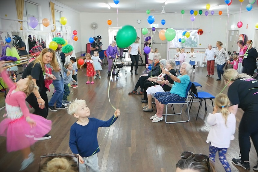 A group of elderly people sit on chairs clapping with young children around them and colourful balloons on the roof.