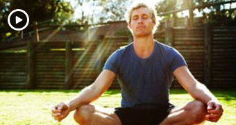 A man sits in a yoga pose on grass in the sunshine.