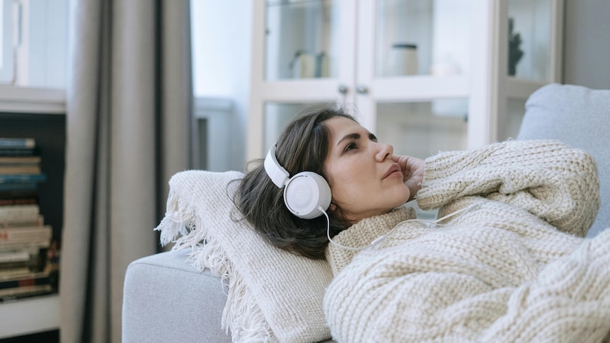 woman in a cream sweater lies on a couch with white headphones on