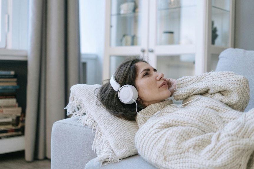 woman in a cream sweater lies on a couch with white headphones on