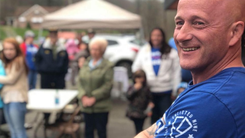 A man wearing a blue T-shirt poses with his arms folded across his chest. Many tattoos are visible on the man's upper arms.