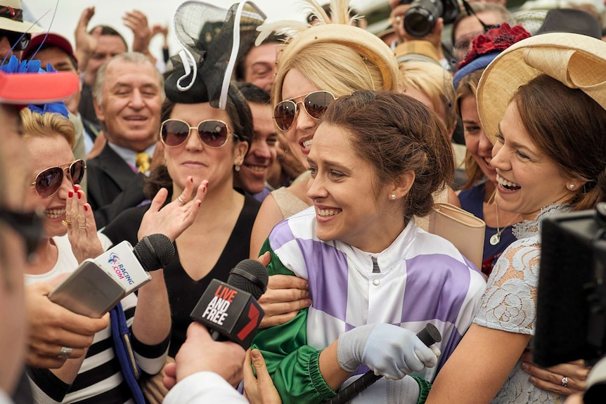 A jubilant female jockey is surrounded by happy people at a horse racing meet.