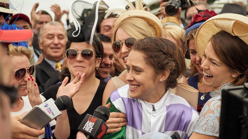A jubilant female jockey is surrounded by happy people at a horse racing meet.