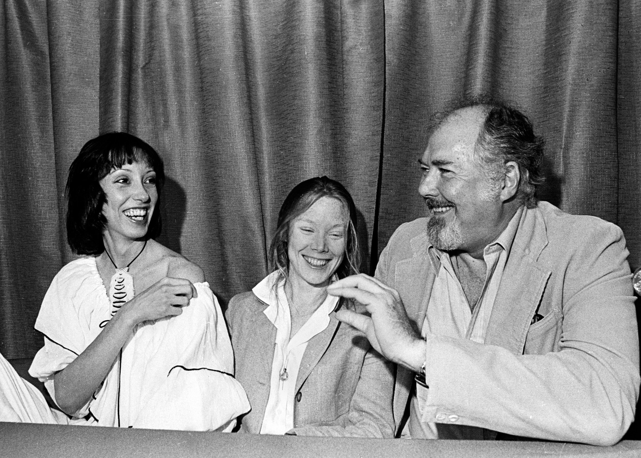 Shelley Duvall, Sissy Spacek and Robert Altman sit at a table, laughing. 