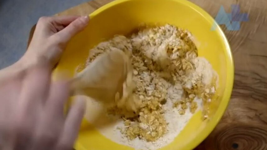 A hand stirs flour and oats in a bowl with a wooden spoon
