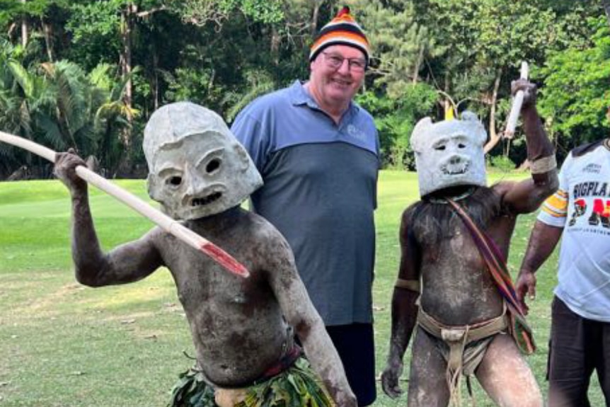 A man stands between two masked men holding spears.