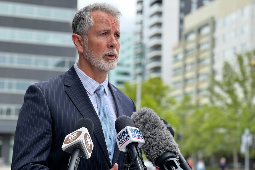 A man in a suit speaks into multiple microphones.