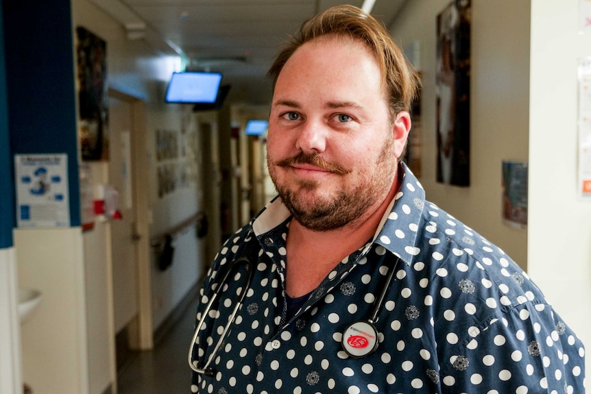 A man with a moustache and beard stands in a hospital hall with a stethoscope around his neck, smiling.