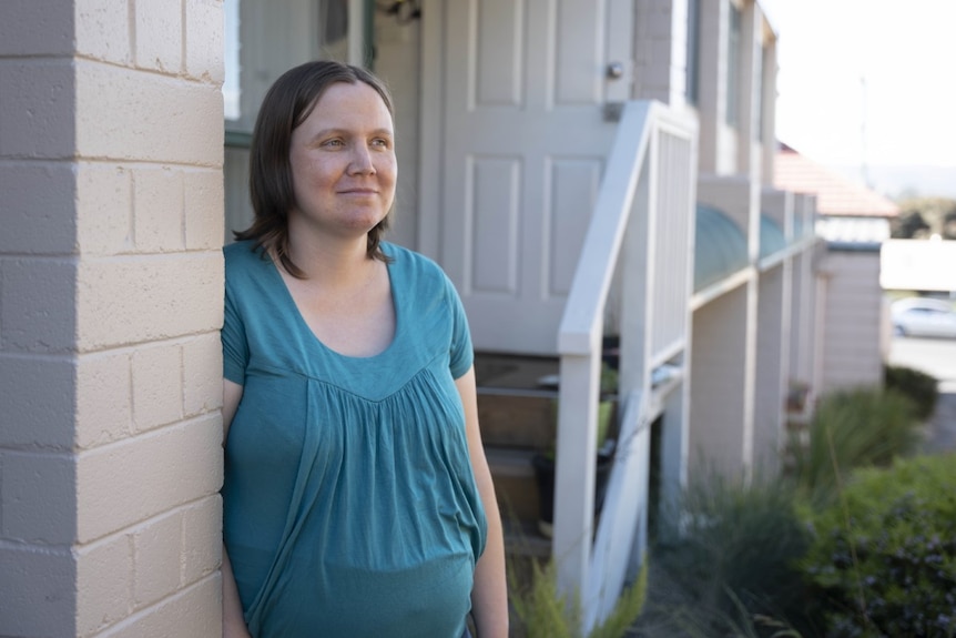 A young pregnant woman with medium length brown hair. She is on a porch, looking off camera