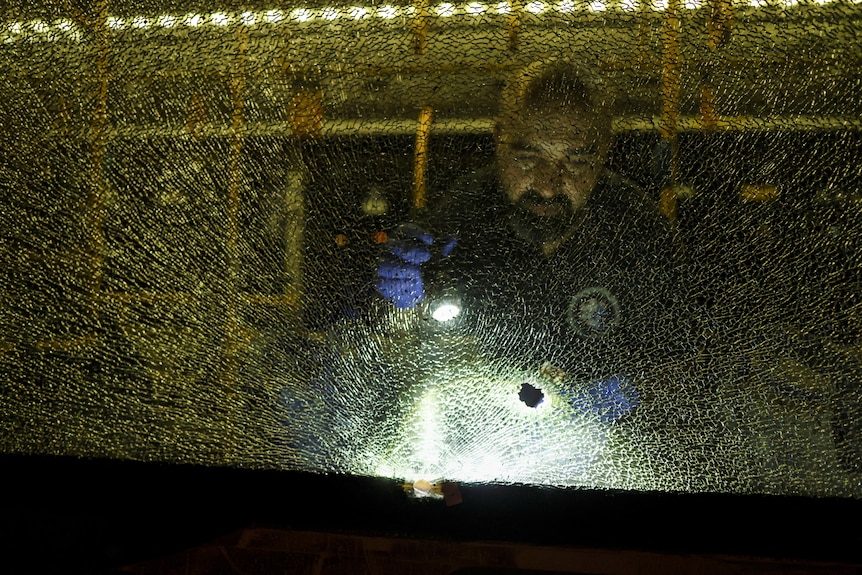 一名秃头侦探站在一辆被砸坏的公共汽车车窗后面，用手电筒检查弹孔。