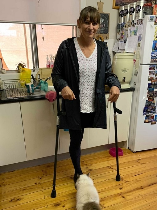 A woman is standing in a kitchen holding herself up with crutches. She has one leg and there is a cat in front of her.
