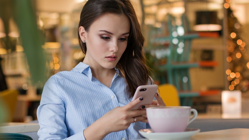 Woman with brown hair sits at a cafe table looking down at her phone, in a story about 'can we talk' texts and anxiety.