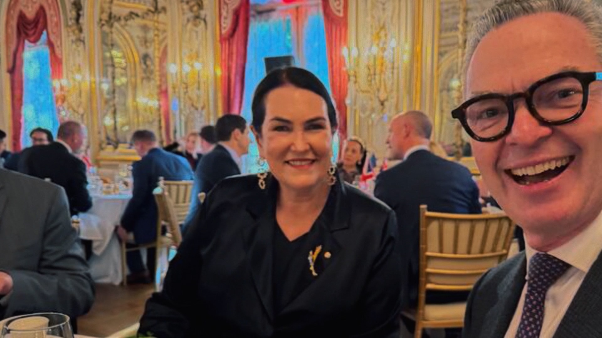 A middle-aged brunette woman wearing all black next to a middle-aged man inside a fancy ballroom.