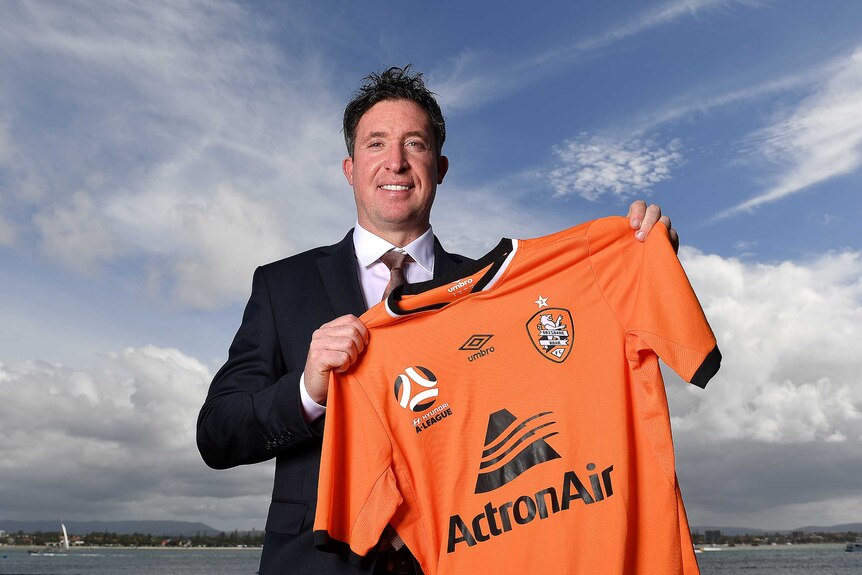 The sky is a resplendent blue as a beaming Robbie Fowler stands on a water front holding aloft an orange Brisbane Roar jersey.