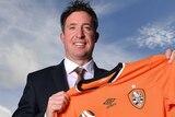 The sky is a resplendent blue as a beaming Robbie Fowler stands on a water front holding aloft an orange Brisbane Roar jersey.