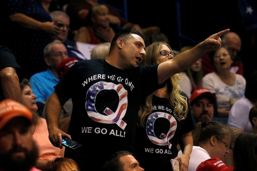 Two people with QAnon t-shirts stand in a crowd at a Trump rally.