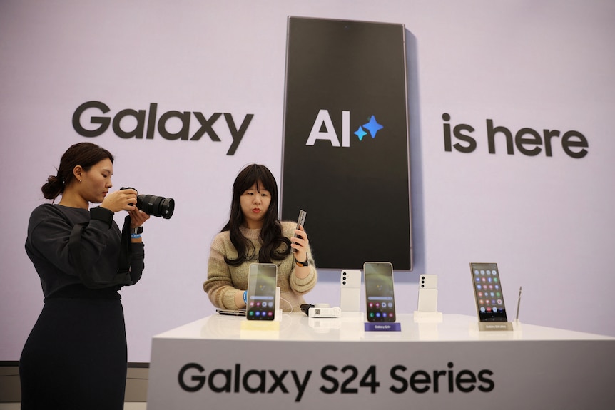 A medium shot of a woman using a camera to photograph another woman who is holding a mobile phone in a Samsung store.