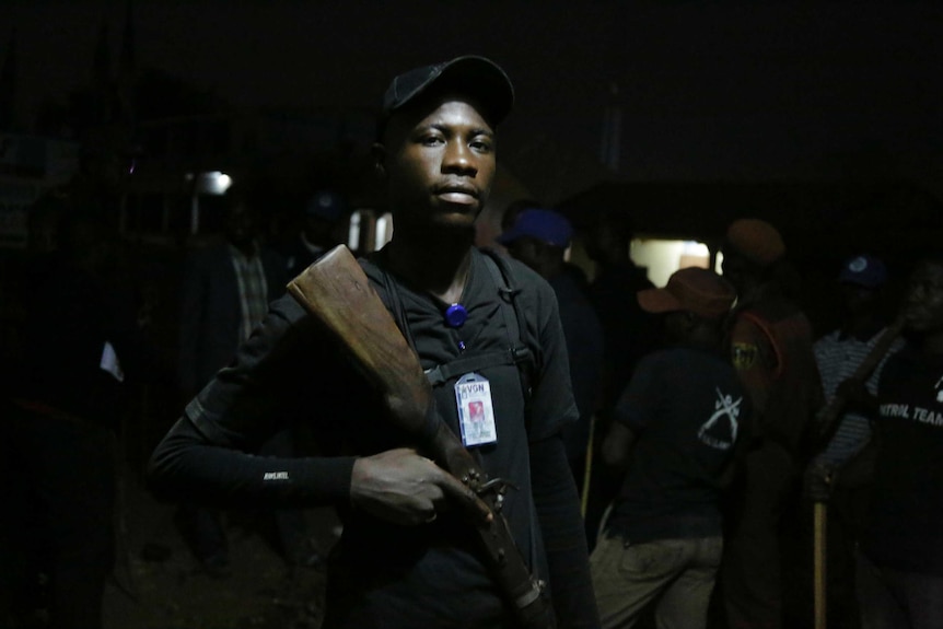 A close up of a vigilante with a black cap, black clothes holding a rifle and staring straight at the camera.