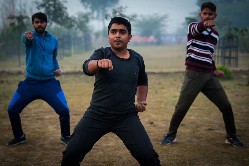 Three men stand in formation, all in the same pose with one fist extended in front and the other at their hip