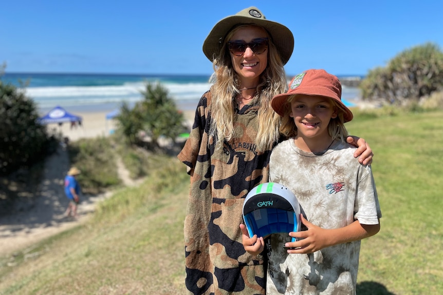 a mother and son standing arm in arm at the beach. the boy is holding a helmet.