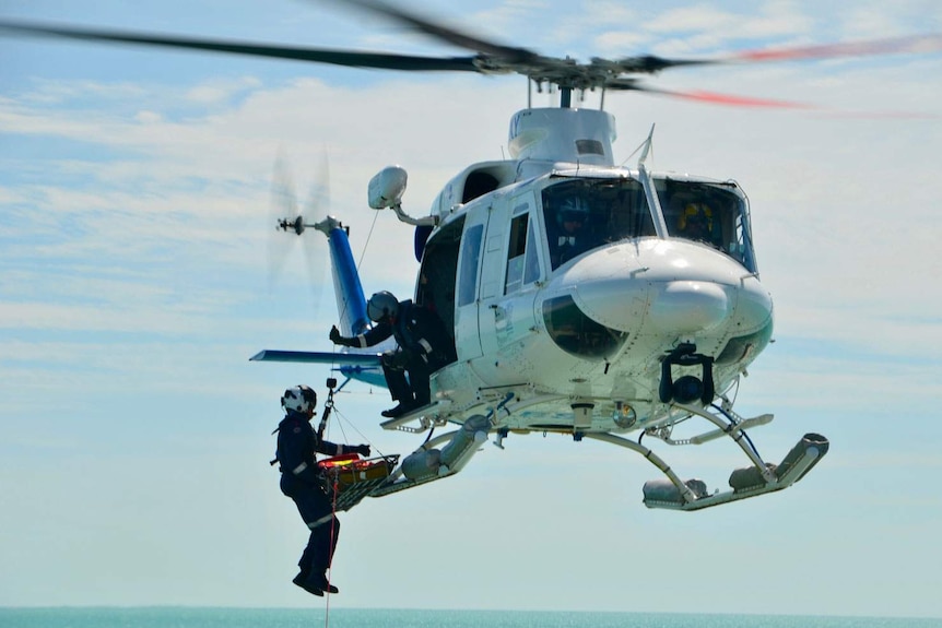 Rescue helicopter in flight over ocean with a crew member being winched with stretcher.