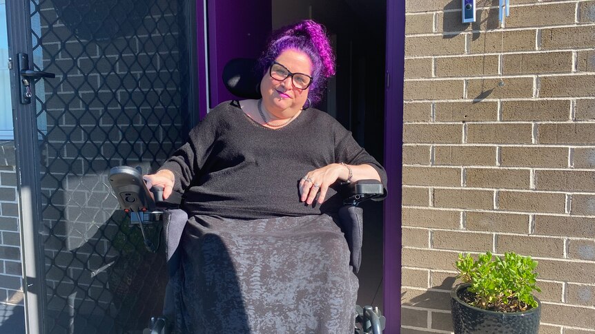 A woman with purple hair, sitting in a wheelchair, in front of her brick house. 