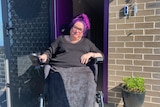A woman with purple hair, sitting in a wheelchair, in front of her brick house. 