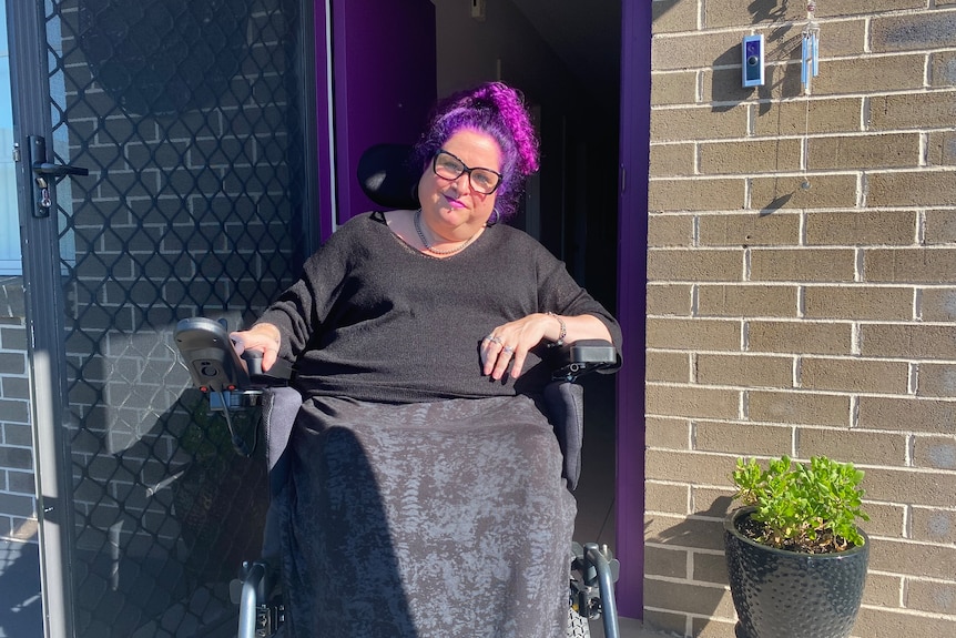 A woman with purple hair, sitting in a wheelchair, in front of her brick house. 