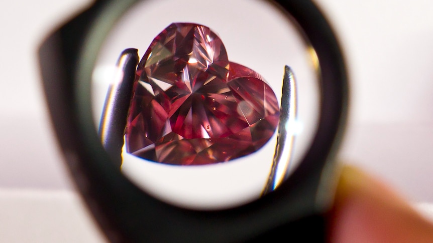 A heart-shaped pink diamond held by tweezers under a magnifying glass