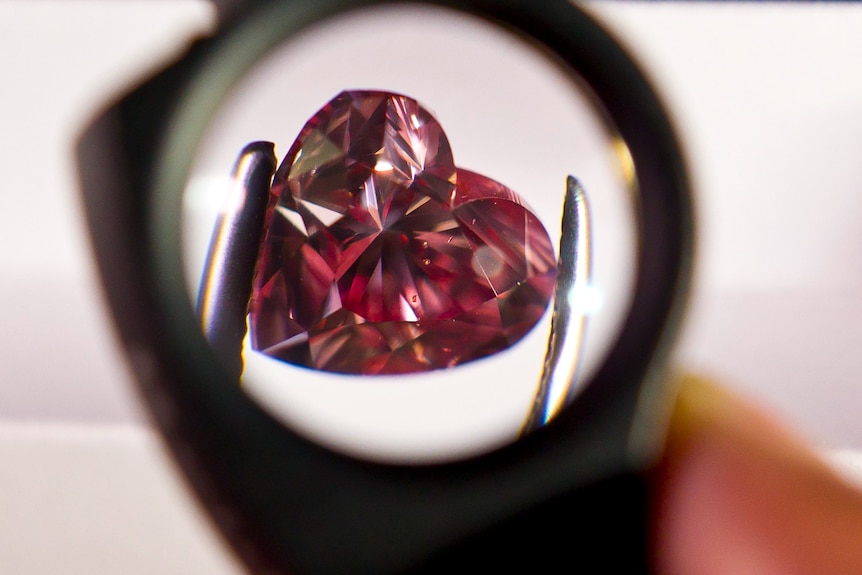 A heart-shaped pink diamond held by tweezers under a magnifying glass