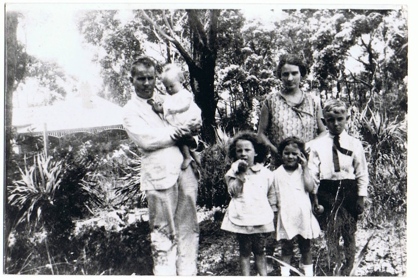 A man in a black and white photo stands outside holding a baby, with a woman and three small children. 