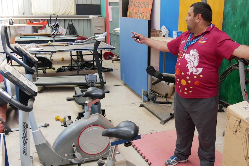A man in a maroon shirt stands with his arms  outspread in front of gym equipment in a warehouse.