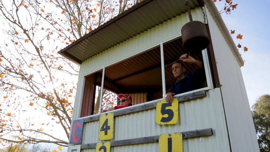 Two men in a scoreboard box look out towards the football ground as one of them rings a scorers bell.