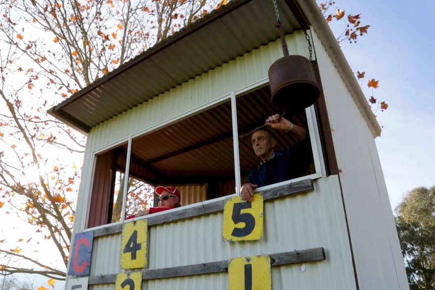 Two men in a scoreboard box look out towards the football ground as one of them rings a scorers bell.