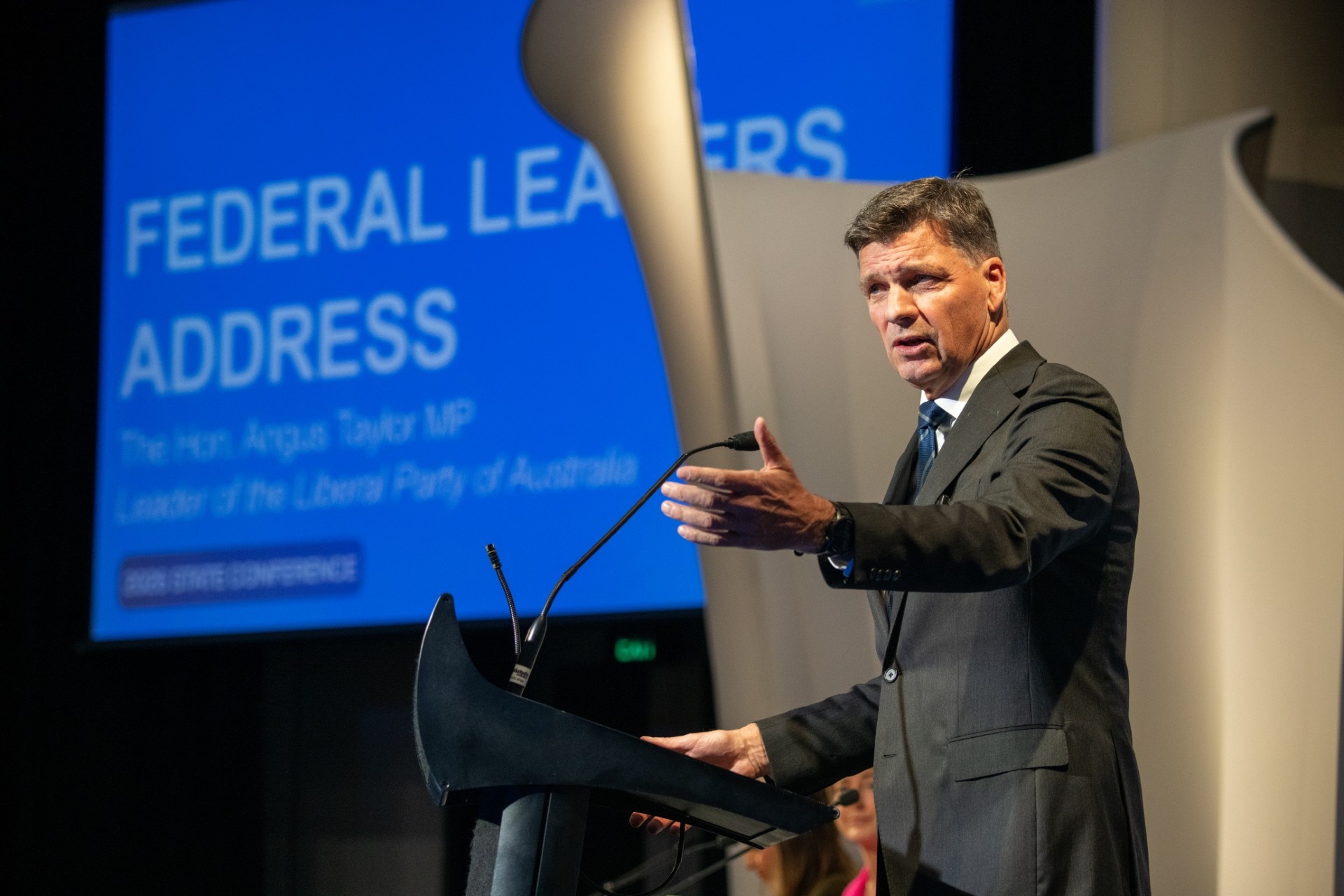 A man in a dark suit and dark hair speaks at a podium with a blue screen behind him.