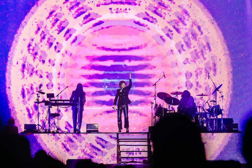 Beck on stage with hand aloft in front of a big screen with a psychedelic pattern on it