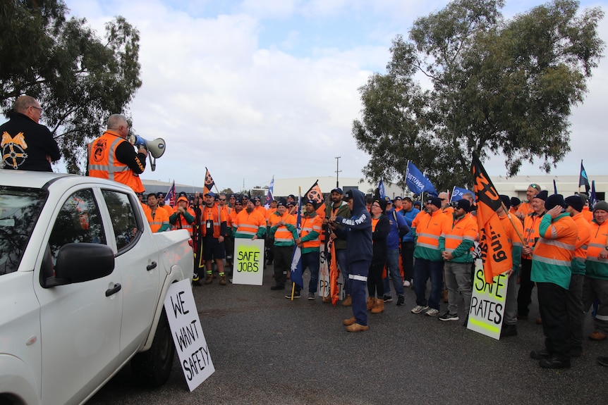 A man with a megaphone on a ute addresses a crown of men wearing high viz 