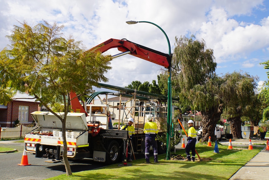 A group of council workers stand next to a truck on a suburban street, that has a machine carrying out work on a lightpole.