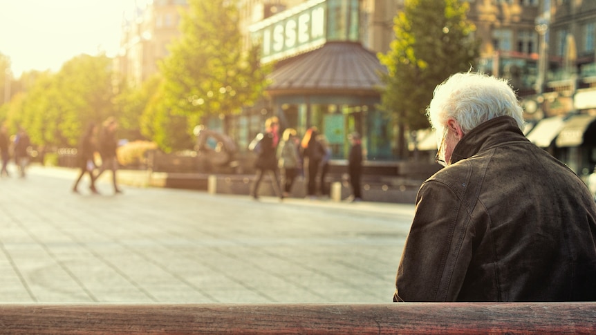 An elderly person in a leather jacket sits on a bench in public and looks down at their lap